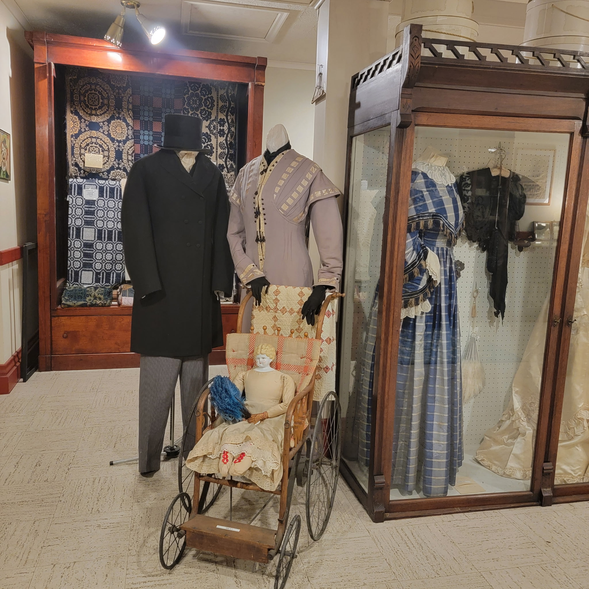 Period costume display with a frock coat, gowns and an antique pram