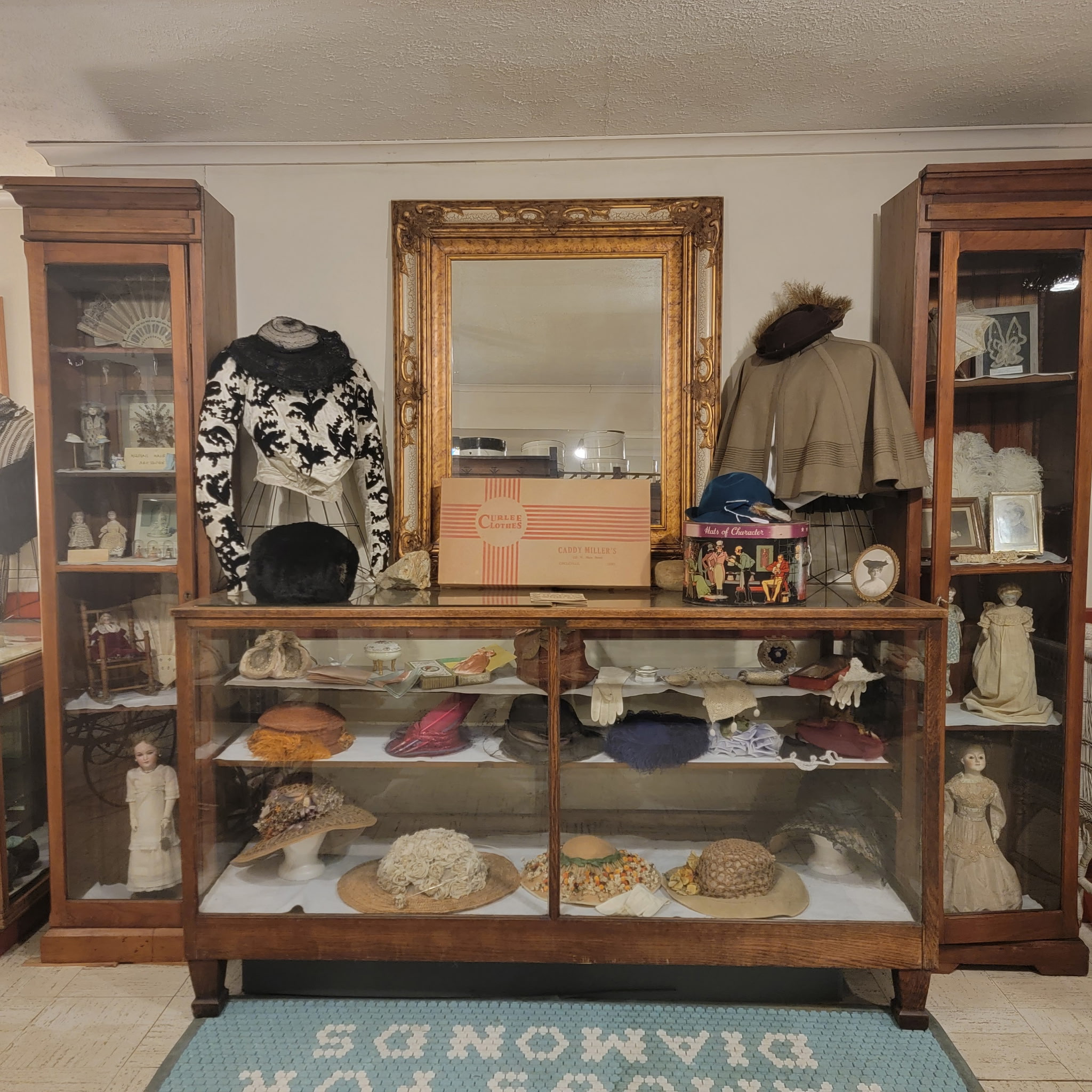Display case of antique hats, gloves and dolls beneath a gilt mirror